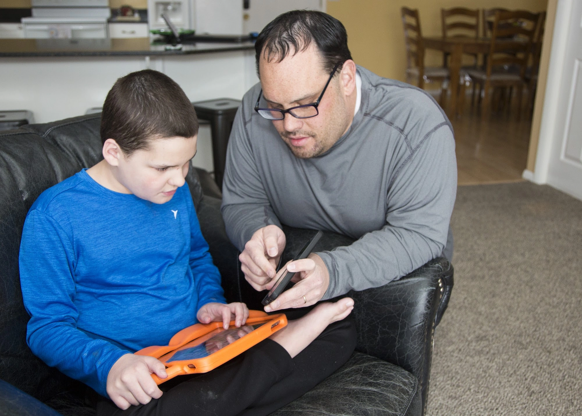 A young AAC user in a blue shirt and an adult look at tablets together on a sofa.